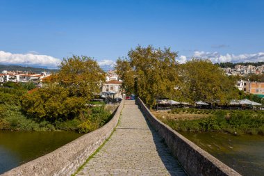 Yunanistan 'ın Arta kentindeki antik taş köprünün kaldırım taşı yolu boyunca, yapraklı nehir kenarındaki ağaçlara ve ötesindeki kasabaya doğru bakın. Parlak gün ışığı, desenli taşları, yeşil yaprakları ve nehrin üzerinde açık mavi gökyüzünü ortaya çıkarıyor..