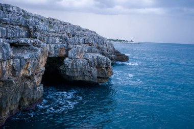 Belirgin bir deniz mağarası ve dokulu kaya oluşumlarıyla, derin mavi Adriyatik Denizi 'nin üzerinde sivri kireçtaşı kayalıkları yükseliyor. Bulutlu gökyüzü ve engebeli kıyı şeridi doğal bir drama ve kıyı vahşeti hissi uyandırıyor..
