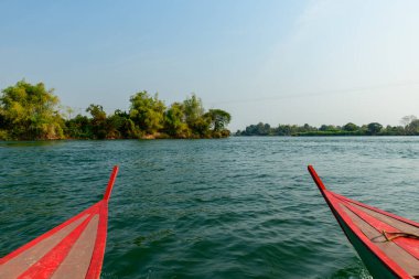 Laos 'un güneyindeki 4000 ada bölgesindeki Mekong Nehri üzerindeki yemyeşil adalara doğru kırmızı çizgili iki tahta tekne pruvası. Berrak gün ışığı, dalgalı suyu ve geniş mavi gökyüzünün altındaki doğal bitki örtüsünü aydınlatır..
