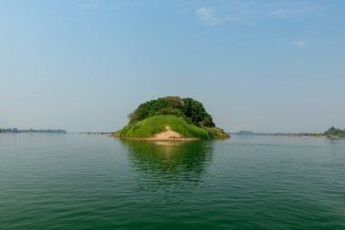 Laos 'un güneyindeki Champassak yakınlarındaki Mekong Nehri' nin sakin sularından yoğun yeşillik ve kumlu kıyılarla kaplı tek bir bereketli ada yükselir. Açık gün ışığı ve geniş mavi bir gökyüzü yumuşak yansımalar ve doğal yansımalarla sakin bir nehir manzarası oluşturur.