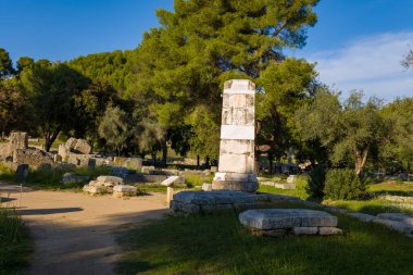 Antik Paios Zaferi anıtının mermer heykeli Yunanistan 'ın Olympia kentindeki dağınık harabeler ve yemyeşil alanlar arasında yer alıyor. Sıcak öğleden sonra güneşi, bu tarihi günde dokulu taşları, etrafındaki çam ve zeytin ağaçlarını vurguluyor.
