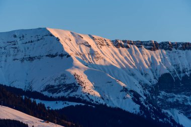 Mont Jolys dramatik kar kaplı tepe sıcak altın saat güneş ışığıyla parıldıyor, çarpıcı dokuları ve Fransız Alpleri 'ndeki karanlık bir çam ormanının üzerindeki derin gölgeleri ortaya çıkarıyor..