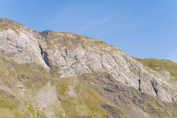 Fransız Pirenesi 'nde Gavarnie Gedre yakınlarında berrak mavi bir gökyüzünün altında kayaların tepeleri ve dağınık çimenlik alanları olan engebeli bir dağ yamacı aydınlatılır. Sahne zıt dokuları ve doğal toprak tonlarını vurgular.