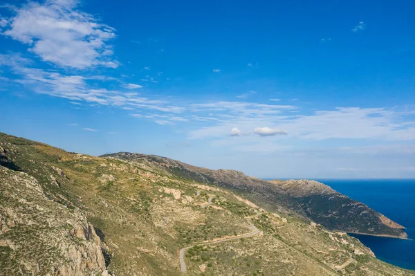 Kapsamlı bir hava manzarası, Yunanistan 'ın Magne kentindeki derin mavi Akdeniz' e bakan dolambaçlı toprak raylı kayalık tepeleri ortaya çıkardı. Sahne aydınlık gün ışığıyla yıkanır ve dağınık bulutlar geniş gökyüzünü vurgular..