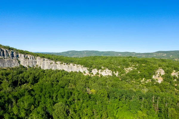 Geniş bir kireçtaşı plato, Gorges de Ardeche 'deki canlı mavi gökyüzünün altında yemyeşil ağaçlardan oluşan yoğun bir gölgeliğin üzerinde uzanır. Sahne, parlak yaz güneşiyle yıkanmış doğal dokuları ve canlı manzarayı vurguluyor..