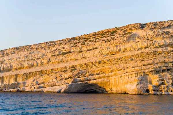 Girit 'in Matala kentindeki mavi denizin üzerinde öğleden sonra güneşiyle aydınlanan katmanlı kireçtaşı kayalıkları yükseliyor. Kayalık oluşumlar desenli yüzeyler ve doğal mağaralar sergiliyor, dramatik bir kıyı manzarası yaratıyor..