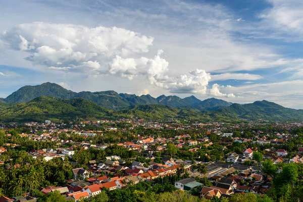 Kuzey Laos 'taki Luang Prabang' a geniş bir manzara, yemyeşil tepeler ve dramatik kümülüs bulutlarıyla çevrili kırmızı çatılardan oluşan bir demet sunar. Canlı tropikal şehir manzarası, parlak bir gökyüzünün altındaki ormanlı dağ zeminine kurulmuştur..