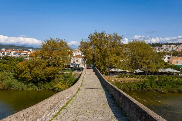Yunanistan 'ın Arta kentindeki antik taş köprünün kaldırım taşı yolu boyunca, yapraklı nehir kenarındaki ağaçlara ve ötesindeki kasabaya doğru bakın. Parlak gün ışığı, desenli taşları, yeşil yaprakları ve nehrin üzerinde açık mavi gökyüzünü ortaya çıkarıyor..