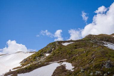 Aiguillette des Houches engebeli yeşil bir tepe sergiliyor. Dalgalı mavi bir gökyüzü ve dağınık kümülüs bulutlarının altında kalan karla benekli..