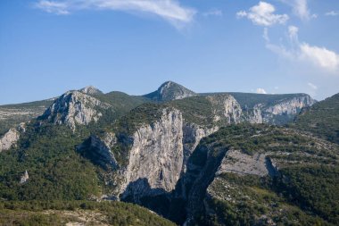 Jagged kireçtaşı uçurumları Verdon Vadisi 'ndeki yoğun yeşil ormanın üzerinde yükseliyor, engebeli tepeler ve ince bulutlarla benekli koyu mavi bir gökyüzü..