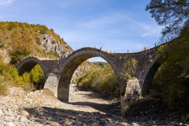 Yunanistan 'ın Epirus kentindeki Kipoi yakınlarında sonbahar yaprakları ve engebeli tepelerle çevrili üç taş kemer ile tarihi Plakidas Köprüsü kayalık nehir yatağını kaplar. Parlak gün ışığı desenli duvarcılığı ve Pindus Dağları 'nın canlı manzarasını gözler önüne seriyor..