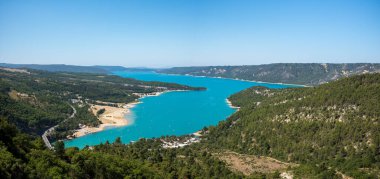 Sainte-Croix gölünün panoramik manzarasında kumlu kıyılarla çevrili canlı turkuaz su ve parlak mavi gökyüzünün altındaki yoğun yeşil tepeler görülüyor..