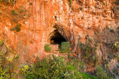 Yığılmış taşlarla çerçevelenmiş engebeli mağara ağzı yemyeşil ve yabani bitkilerle çevrili canlı turuncu kireçtaşı bir uçurumda açılır. Sıcak güneş ışığı kayalık arazinin toprak dokularını ve doğal formlarını güçlendirir..