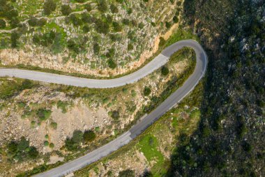 Yunanistan 'ın Vathia kenti yakınlarındaki engebeli kayalık arazide kıvrılan dar bir dağ yolunda keskin bir eğrinin havadan görünüşü. Güneş ışığı, dokulu kayalıkları ve doğal manzarayı vurgular.