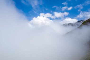 Col du Bonhomme 'da kısmen gizlenmiş dağ yamaçlarında yoğun beyaz sis kıvrılır. Parlak mavi gökyüzü ve dağılmış bulutlar gizem ve yükseklik hissi yaratır..