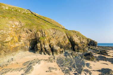 Normandiya 'nın Cap de Carteret kentindeki engebeli kumlu plajın üzerinde yeşil bitki örtüsü olan dik kayalıklar yükseliyor. Ilık güneş ışığı, berrak mavi gökyüzü ve sakin denizle çelişen kayaları vurgular..