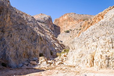 Dik, engebeli kaya duvarları Girit 'teki Seitan Limania' da kurak, taştan bir vadide yükseliyor. Sıcak güneş ışığı kireç taşının dokusunu aydınlatıyor ve açık mavi gökyüzünün altındaki seyrek bitki örtüsünü aydınlatıyor..