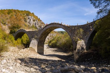 Tarihsel üçlü kemerli Plakidas Köprüsü, Yunanistan 'ın Epirus kentindeki Kipoi yakınlarında sonbahar yaprakları ve engebeli tepelerle çevrili kuru, taşlı bir nehir yatağını kaplıyor. Parlak gün ışığı desenli taş işçiliğini ve Pindus Dağları 'nın doğal manzarasını gözler önüne seriyor..