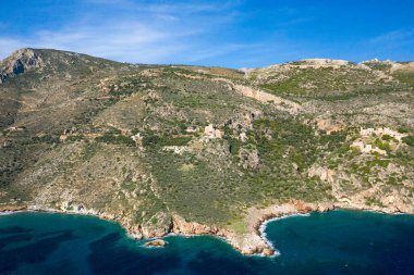 Yunanistan 'ın Mani kentindeki berrak turkuaz su ve kayalık kıyı üzerindeki engebeli bir yamaçta kurulmuş dağınık bir taş yerleşiminin hava manzarası, parlak mavi bir gökyüzünün altında.