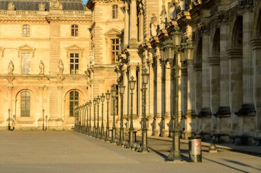 Erken güneş ışığı, Fransa 'nın başkenti Paris' teki Louvre Sarayı 'nın tarihi cephesi boyunca bir sıra süslemeli sokak lambası ve klasik sütunları aydınlatır. Sıcak ışık simetriyi, heykeli ve detaylı rönesans taş işçiliğini geliştirir..