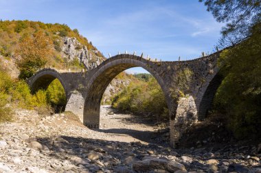 Tarihsel üçlü kemerli taş köprü, Yunanistan 'ın Epirus kentindeki Kipoi kenti yakınlarında sonbahar yaprakları ve engebeli tepelerle çevrili kuru bir kayalık nehir yatağını kaplıyor. Parlak gün ışığı, Pindus Dağları 'nın dokulu ve canlı doğal manzarasını vurgular..