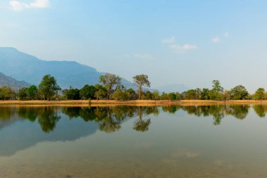 Dağılmış ağaçların yansımaları Güney Laos 'taki Wat Phu yakınlarında sakin bir göletin kenarında. Sıcak gün ışığı ve uzak dağ yamaçları yumuşak doğal tonlarla ve açık gökyüzüyle huzurlu bir manzara yaratır..