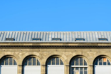 Simetrik kemerli pencereleri ve çinko çatısı olan tarihi bir taş binanın üst kısmı. Parlak güneş ışığı altında parlak mavi gökyüzüne karşı kurulmuş..