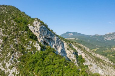 Görülebilir mağaraları olan sivri kireçtaşı kayalıkları, açık mavi yaz gökyüzü ve uzak dağ sırtlarının altında, Verdon Boğazı 'ndaki yemyeşil ormanın üzerinde yükseliyor..