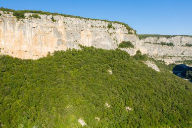Gorges de Ardeche 'deki yoğun ve canlı bir ormanın üzerinde yükselen yüksek kireçtaşı uçurumlarının havadan görünüşü. Parlak güneş ışığı ve berrak mavi gökyüzü engebeli kaya dokusunu ve yemyeşil yaprakları güçlendirir..
