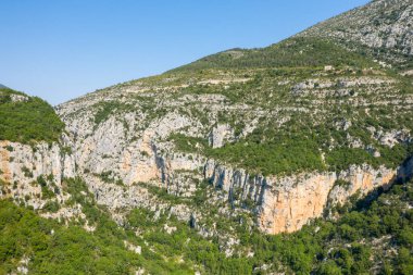 Yoğun yeşil bitki örtüsüyle kaplı geniş kireçtaşı terasları açık mavi gökyüzü altında parlak yaz güneşiyle aydınlatılan Verdon Boğazı 'nın dramatik uçurumlarının üzerinde yükseliyor..