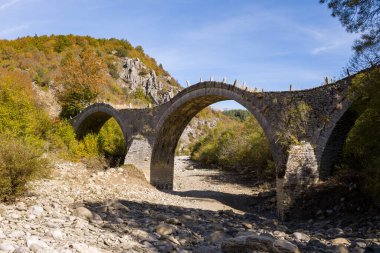 Üç kemerli Plakidas Köprüsü, Yunanistan 'ın Epirus kenti Kipoi yakınlarındaki renkli sonbahar yaprakları ve engebeli tepelerle çevrili kuru bir kayalık nehir yatağını kaplar. Parlak gün ışığı Pindus Dağları 'nın dokulu duvarcılığını ve canlı manzarasını ortaya çıkarır..