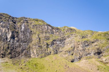 Dikey kaya duvarları, Pireneler 'in dağlarındaki eğimli otlak bir tepenin üzerinde berrak mavi gökyüzünün altında güçlü bir gün ışığında yıkanır. Engebeli dokular ve zıt toprak renkleri çarpıcı bir alp manzarası yaratır..
