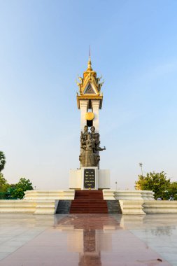 Kamboçya 'nın Phnom Penh kentindeki Dostluk Anıtı' nın dikey görüntüsünde altın kuleye sahip uzun bir sütunun temelinde heykel grubu yer alıyor. Anıt, açık bir plaza ve açık bir gökyüzünün altındaki yeşilliklerle çevrili bir platformda duruyor..