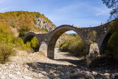 Yunanistan 'ın Epirus kentindeki Kipoi yakınlarında sonbahar yaprakları ve engebeli tepelerle çevrili üç taş kemeriyle tarihi Plakidas Köprüsü, kayalık nehir yatağını kaplamaktadır. Parlak gün ışığı, PIndus 'in dokulu ve canlı doğal yapısını vurgular.