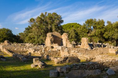 Yunanistan 'ın Olympia kentindeki Güney Hamamları' nın antik Roma tuğlaları ve taş duvarları olgun zeytin ağaçları ve yemyeşil alanlar arasında yer alıyor. Sıcak öğleden sonra güneş ışığı, berrak mavi gökyüzü altında dokulu ve sakin arkeolojik manzarayı vurgular..