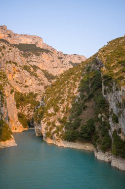 Yumuşak sabah ışığı, dar kireçtaşı kanyon duvarlarını ve Verdon Boğazı 'ndaki turkuaz suyunu, engebeli dokuları ve berrak bir gökyüzünün altında dağınık yeşil bitki örtüsünü ön plana çıkarıyor..