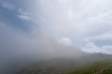Uçsuz bucaksız bir alp zirvesi, bulutların arasından süzülen yumuşak güneş ışığıyla kısmen gizlidir ve solgun bir gökyüzünün altındaki kayalık araziyi ortaya çıkarır..
