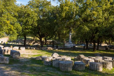 Yunanistan 'ın Olympia arkeoloji sahasındaki olgun meşe ağaçlarının altında dağılmış antik taş bloklar bulunuyor. Sıcak öğleden sonra güneş ışığı, bu tarihi kalıntıları çevreleyen dokulu taş ve doğal yeşilliği vurguluyor..