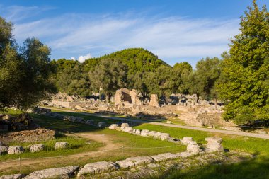 Güney hamamlarının antik Roma tuğla ve taş duvarları Yunanistan 'ın Olympia kentinde dağınık duvarlar ve yemyeşil yeşillikler arasında duruyor. Sıcak öğleden sonra güneş ışığı, sık ormanlık tepeler ve açık mavi gökyüzü ile dokulu harabeleri vurguluyor..