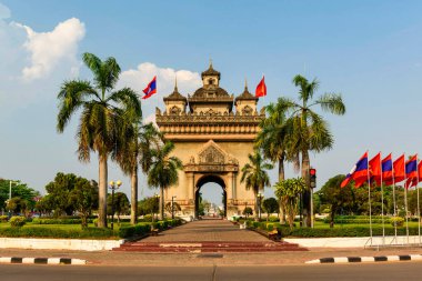 Patuxai anıtı, merkezi Vientiane, Laos 'ta, palmiye ağaçları, manikürlü bahçeler ve ulusal bayraklarla çerçevelenmiş olarak duruyor. Karmaşık mimari detaylar ve geleneksel Lao motifleri net gün ışığında vurgulanarak dengeli bir