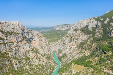 Verdon Vadisi 'ni saran canlı turkuaz bir nehrin üzerinde yükselen sarp kireçtaşı kayalıkların geniş hava manzarası. Berrak mavi bir gökyüzünün altındaki bereketli orman ve güneşli tepelerle çevrili..