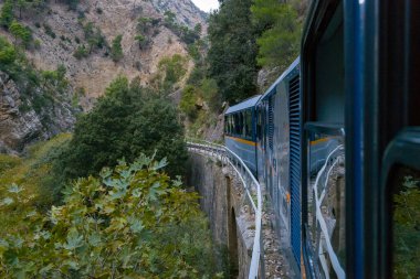 Yunanistan 'ın Kalavrita kenti yakınlarındaki Vouraikos Vadisi' nin yemyeşil ve engebeli uçurumları boyunca eğri bir taş köprü boyunca giden mavi dar çaplı bir tren. Sahnede yoğun yeşillikler, kayalık yamaçlar ve Mora 'nın dramatik manzarası yer alıyor.