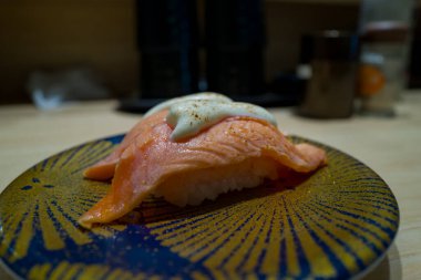Süslü mavi ve altın bir tabakta, hafif aydınlatılmış bir restoranın iç kısımlarında hafif ahşap bir tezgahın üzerine yerleştirilmiş bir parça kremalı beyaz sosla kaplı bir parça somon balığı sunulur..