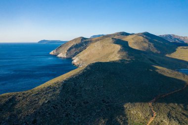 Yunanistan 'ın Cap Tenare kentindeki güneşli tepelerin ve engebeli kıyı şeridinin havadan perspektifi. Açık mavi gökyüzü ve derin gölgeler Akdeniz kıyısındaki dramatik araziyi vurguluyor.