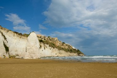 İtalya 'nın Vieste plajında dramatik sahil kayalıkları ve turkuaz Adriyatik Denizi' ne karşı duran uzun beyaz kireçtaşı bir kireç taşı. Sahne, parçalı bulutlu bir gökyüzünün altında hafif gün ışığıyla aydınlatılır.
