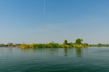 Laos 'un güneyindeki 4000 ada bölgesindeki Mekong Nehri' nin sakin sularından yoğun yeşilliklerle kaplı yemyeşil bir ada yükselir. Parlak gün ışığı ve geniş mavi bir gökyüzü yumuşak yansımalar ve tropikal bir nehir manzarası yaratır.