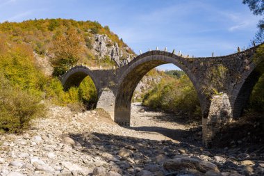 Tarihsel üçlü kemerli Plakidas Köprüsü, Yunanistan 'ın Epirus kentindeki Kipoi yakınlarında sonbahar yaprakları ve engebeli tepelerle çevrili kuru, taşlı bir nehir yatağını kaplıyor. Parlak gün ışığı desenli taş işçiliğini ve Pindus Dağları 'nın doğal manzarasını gözler önüne seriyor..