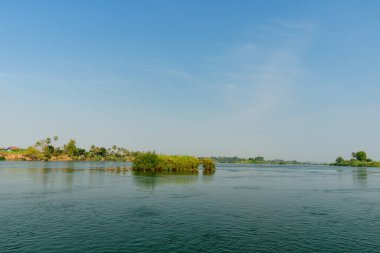 Laos 'un güneyindeki 4000 adanın Mekong Nehri' nin geniş manzarası, küçük yeşil bir ada ve uzak palmiye çizgili nehir kıyıları içerir. Berrak gün ışığı, sakin bir su ve geniş mavi bir gökyüzü, açık alan ve sükunet hissi uyandırıyor..