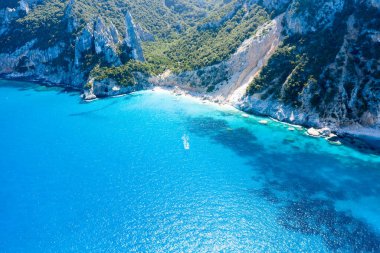 Canlı mavi Akdeniz suları, Cala Goloritze, Baunei, Sardunya 'da dramatik kireçtaşı uçurumları ve yemyeşil alanlarla buluşur. Güneş ışığı denizde parıldıyor, engebeli kıyı şeridini ve doğal dokuları aydınlatıyor..