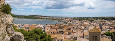 Gruissan köyünün kireçtaşlı kayalar ve yemyeşil yeşilliklerle çerçevelenmiş sakin bir lagünü ve uzak kıyı şeridi olan koyu kireçlenmiş çatılarının panoramik manzarası. Güneş ışığı ve dağınık bulutlar canlı, yaz mevsiminde bir Akdeniz havası yaratır..